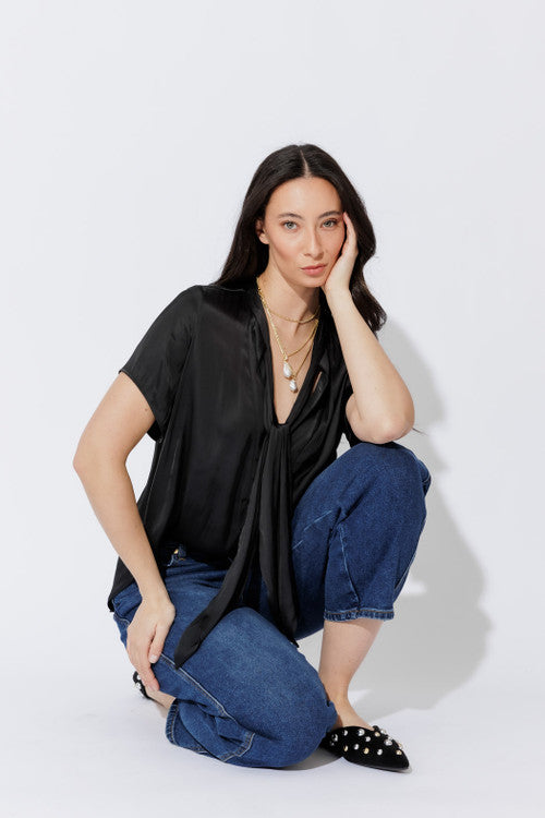 Black silky tie-collar shirt with cuffed denim and studded flats, crouched pose revealing the relaxed drape and V-neckline.