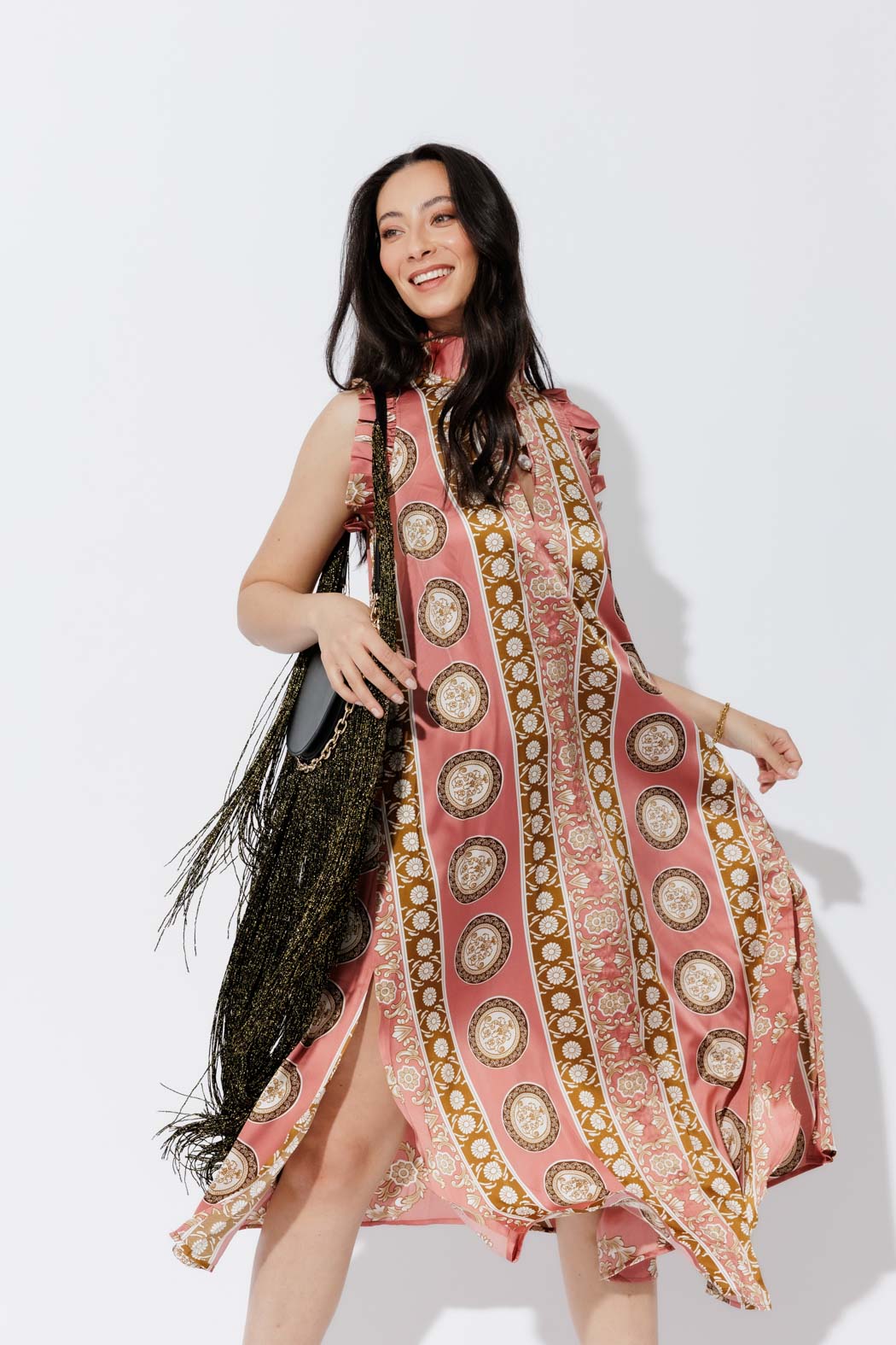 Coral Ruffle Maxi Dress in a flowing A-line silhouette, gold medallion print and ruffle neckline, styled with a fringed bag.