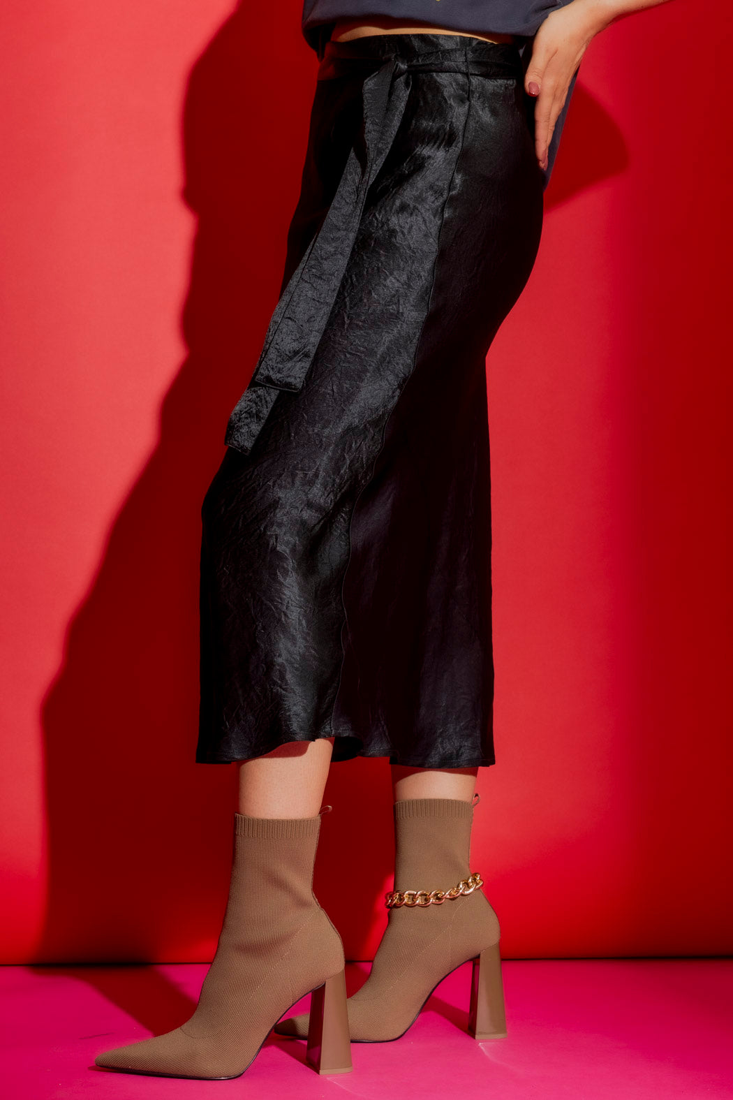 Black Bias Belted Skirt in lustrous satin, styled with tan sock boots against a vivid red backdrop.