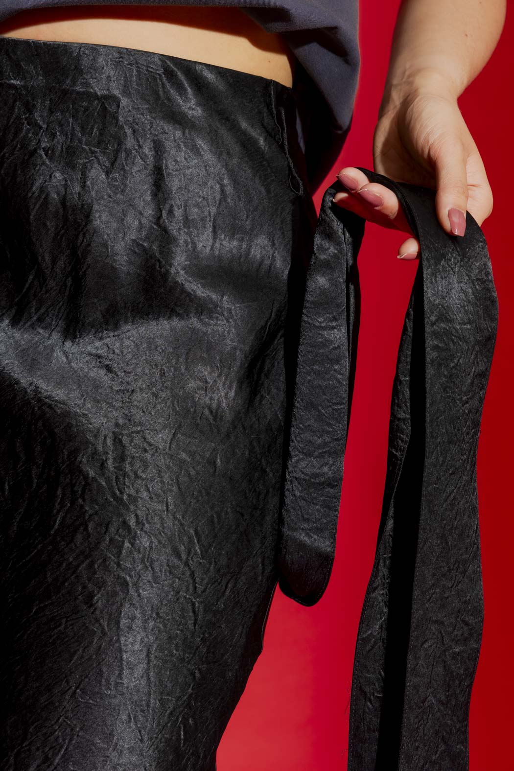 Close-up of the Black Bias Belted Skirt's crinkled satin and self-fabric tie belt, set against a bold red backdrop.