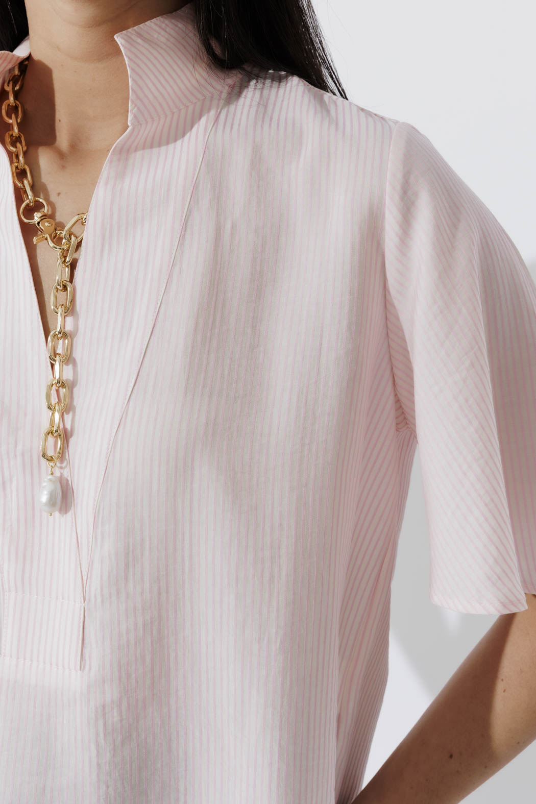 Detail of the Pink Pinstripe Fluted Blogger Shirt — stand collar, V-neck and short sleeve in soft cotton, with a gold chain.