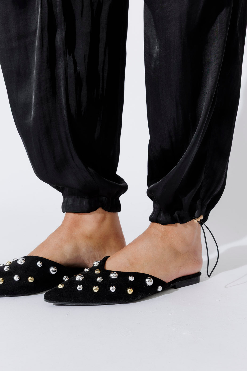 Elevated close-up of the Black Glow Shell Pant's ankle cuff, toggle drawstring and silky sheen, styled with studded mules.